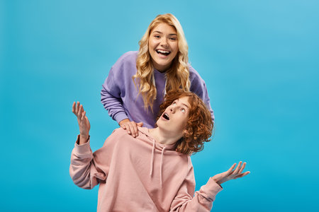excited blonde teen girl having fun and laughing at camera near surprised redhead boyfriend on blue