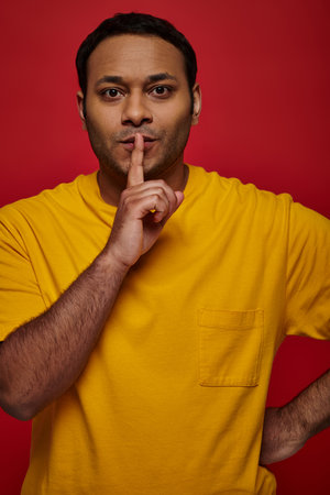indian man in yellow clothes showing hush sign and looking at camera on red backdrop, secretの写真素材