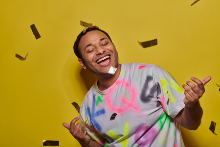 excited indian man in t-shirt gesturing near falling confetti on yellow backdrop, party concept