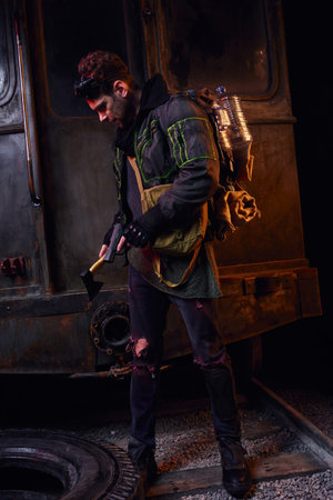 man armed with gun and axe standing with backpack and water bottle in dark abandoned underground