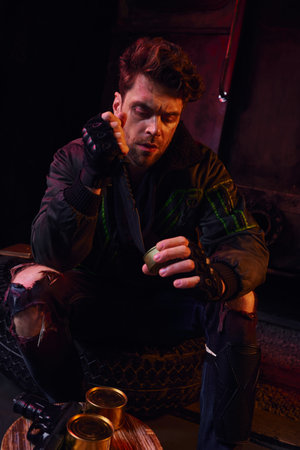 hungry man in worn outfit opening canned food with military knife in dark post-disaster underground