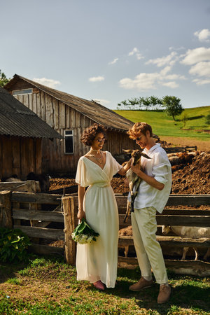 happy multiethnic couple in wedding gown and sunglasses cuddling cute baby goat, countrysideの写真素材