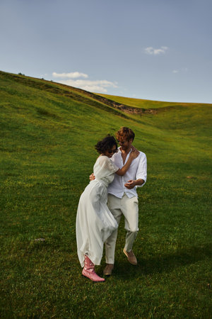 happy newlyweds, asian bride in white dress and cowboy boots walking with groom in green fieldの写真素材