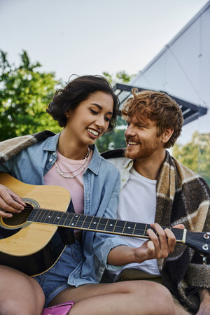 joyful asian woman playing acoustic guitar to happy boyfriend near glass house in countrysideの写真素材