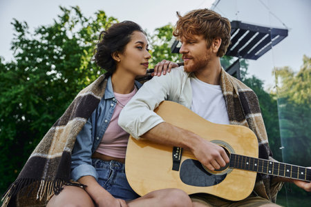 young redhead man playing acoustic guitar to asian girlfriend near glass house, countryside date