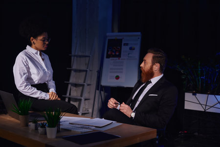 bearded businessman looking at pretty african american woman sitting on work desk at night in office