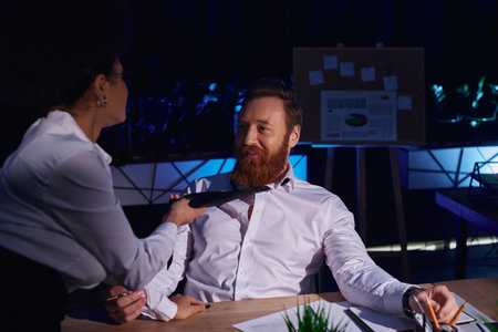 young african american businesswoman pulling tie of happy businessman at workplace in dark office
