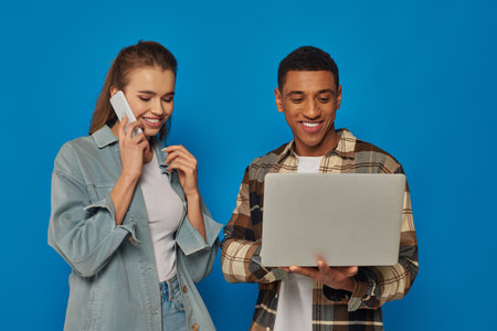 happy african american freelancer using laptop near happy woman talking on smartphone, blue backdrop