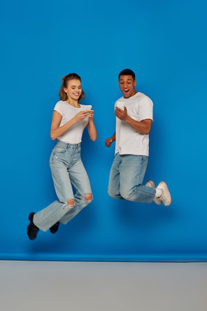 happy multicultural couple using smartphones and jumping  on blue background, full length view