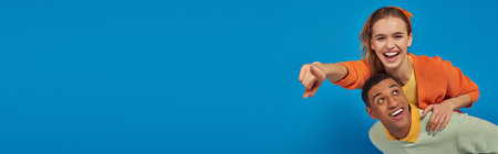 banner, joyful african american man piggybacking girlfriend on blue background, pointing at cameraの写真素材