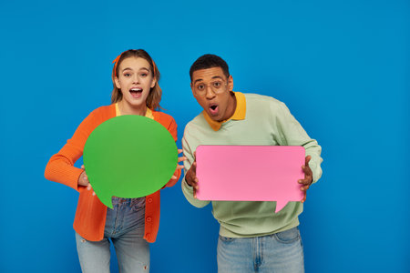excited interracial couple in smart casual clothes holding blank speech bubbles on blue backdrop