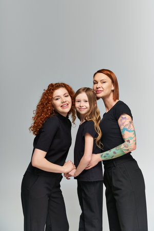 three generations concept, happy redhead women and girl in matching attire hugging on grey backdropの写真素材