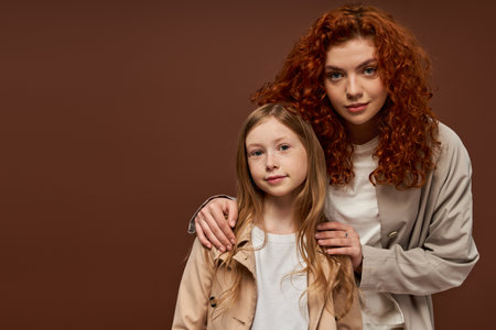 two generations, young curly mother with red hair looking at camera with daughter on brown backdropの写真素材