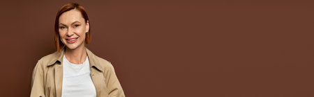 happy redhead woman in beige trench coat looking at camera on brown backdrop, autumn fashion, bannerの写真素材