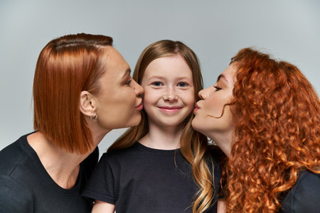 female generations concept, redhead women kissing cheeks of freckled girl on grey backgroundの写真素材
