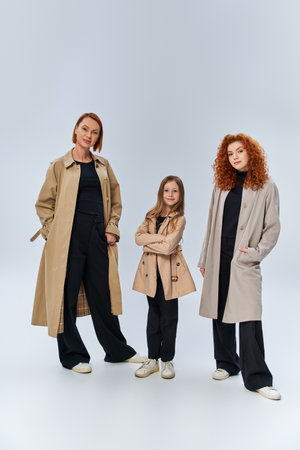 redhead family in coats posing with hands in pockets on grey background, happy female generationsの写真素材
