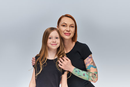 happy woman with tattoo hugging redhead girl and looking at camera on grey backdrop, generationsの写真素材