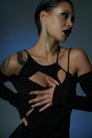 tattooed woman in black stylish dress and sinister makeup looking away on blue and grey backdropの写真素材