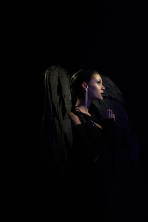 side view of woman in costume of fallen angel with dark wings praying in darkness, black backdropの写真素材