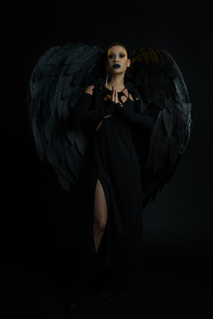 mysterious woman in costume of winged creature standing with praying hands on black, demonic beautyの写真素材