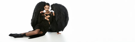 sexy woman in dark makeup and black costume of winged demon sitting on white, bannerの写真素材
