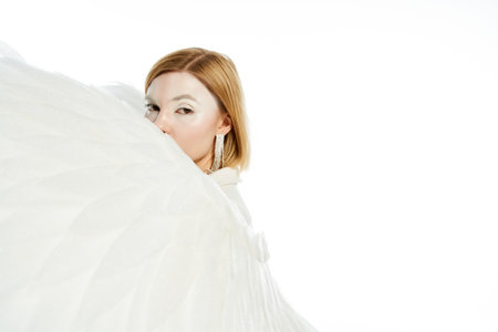 angelic woman looking at camera behind heavenly wings on white backdrop, ethereal beautyの写真素材