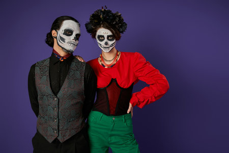 elegant couple in traditional dia de los muertos skeleton makeup looking at camera on blue backdrop