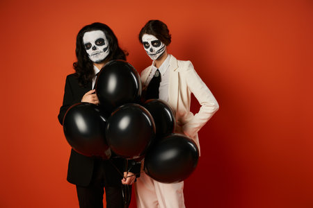 eerie couple in catrina makeup and suits posing with black balloons on red, dia de los muertos partyの写真素材