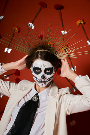 woman in sugar skull makeup and white suit adjusting festive crown on red backdrop with flowersの写真素材