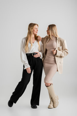 attractive stylish sisters looking at each other and posing together on grey background, fashionの写真素材