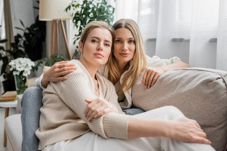 portrait of pretty lovely sisters sitting on sofa hugging and looking at camera, family bondingの写真素材