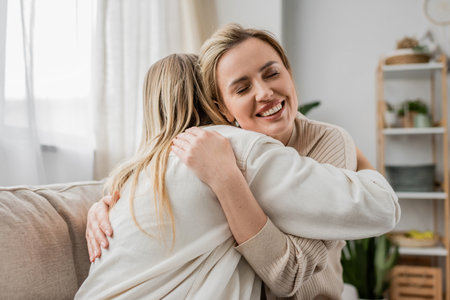 two blonde sisters in lovely casual attire hugging on sofa with closed eyes, family bondingの写真素材