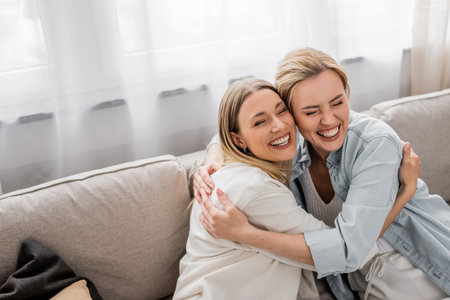 happy sisters in casual outfits sitting on sofa hugging and smiling, quality time, family bondingの写真素材