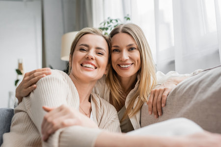 two cheerful trendy sisters in pastel cardigans smiling sincerely at camera, family bondingの写真素材