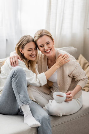 two good looking pretty sisters in trendy attire sitting on sofa and hugging, family bondingの写真素材