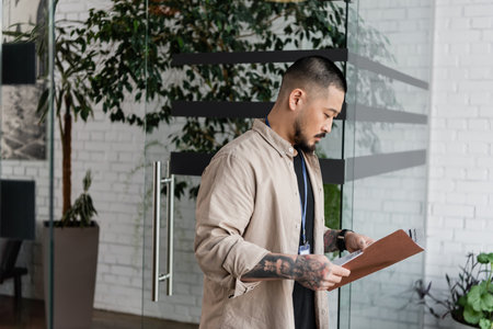 focused asian businessman with tattoo looking at startup plan inside of folder near glass door