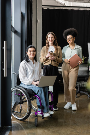 three interracial businesswomen, happy disabled woman in wheelchair working with female teamの写真素材