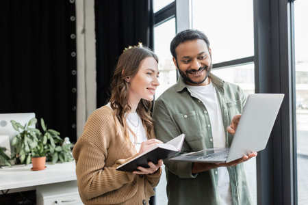 happy indian businessman showing project on laptop to female coworker with notebook, mentorshipの写真素材