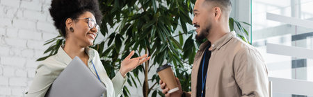 happy african american woman holding coffee and laptop while talking to asian coworker, bannerの写真素材