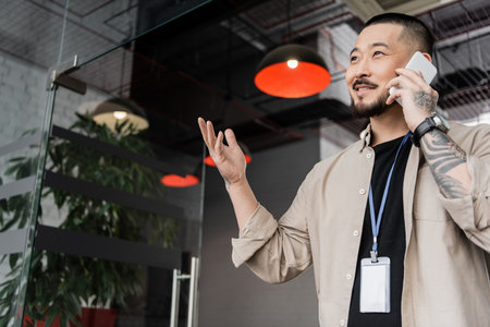 happy asian businessman with tattoo gesturing during phone call smartphone in modern officeの写真素材