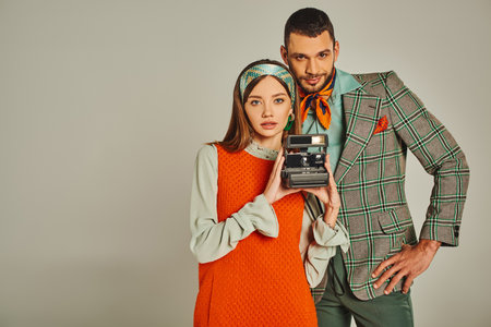 young couple in stylish attire with vintage camera looking at camera on grey, retro-inspired fashionの写真素材