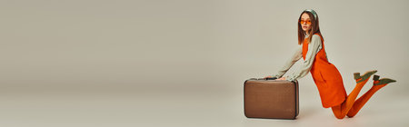 young woman in stylish retro outfit and sunglasses kneeling near vintage suitcase on grey, bannerの写真素材