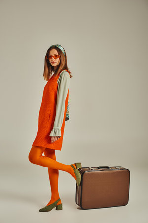 trendy woman in orange dress and sunglasses posing near vintage suitcase on grey, retro styleの写真素材