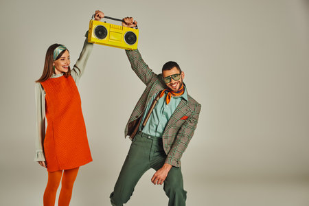 excited couple in stylish vintage clothes holding yellow boombox and dancing on grey backdropの写真素材