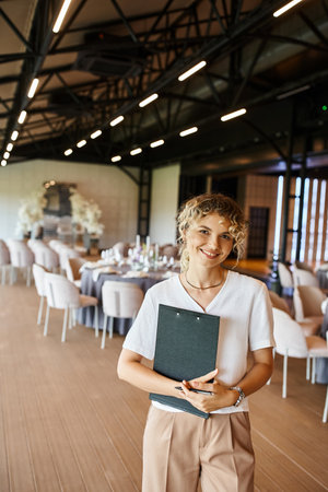 cheerful event manager with clipboard looking at camera near decorated tables in banquet hallの写真素材