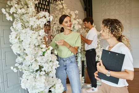 young smiling florist showing white floral decor to event manager with clipboard in event hallの写真素材