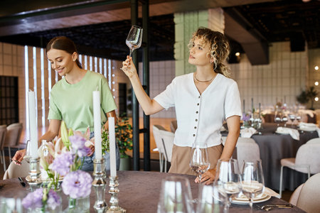 smiling woman looking at clean glass near young colleague decorating festive table in event hallの写真素材