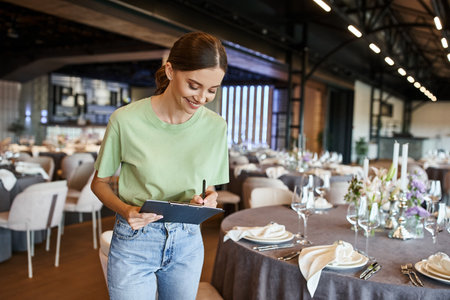 joyful event manager writing notes on clipboard near tables with festive setting in banquet hallの写真素材