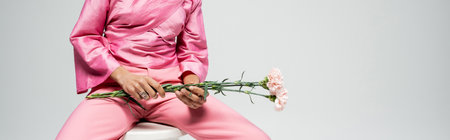cropped fashion model in pink attire holding flowers and sitting on chair, grey backdrop bannerの写真素材