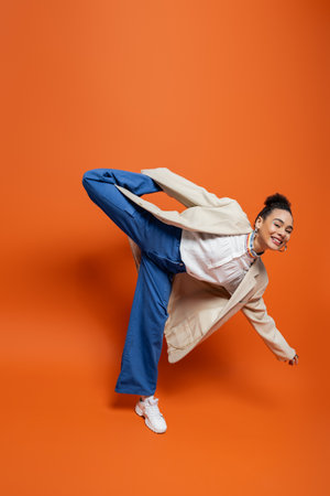 jolly african american model in stylish urban outfit holding playfully raised leg behind her backの写真素材